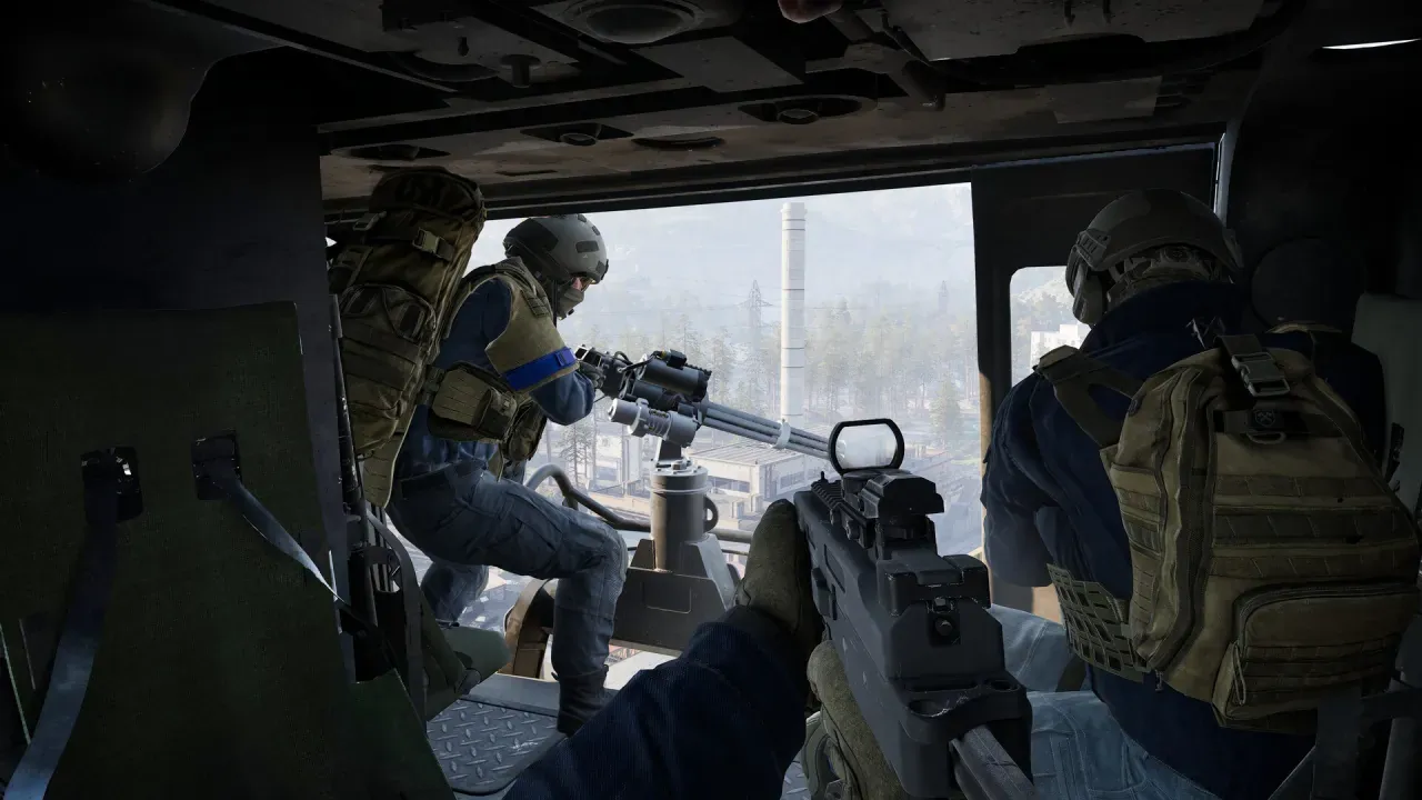 A soldier mans a mounted minigun in a helicopter's open door, above an industrial town with a tall chimney.