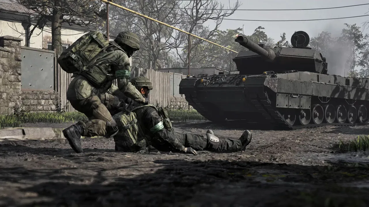 A soldier kneels beside a downed soldier on a muddy village street, with a tank parked behind them.